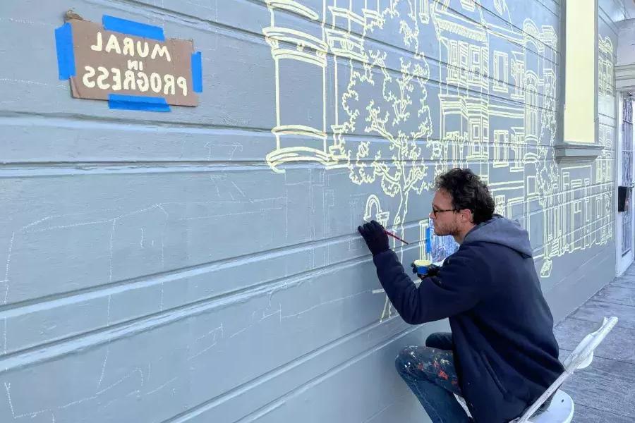 An artist paints a mural on the side of a building in the Mission District, 在建筑上贴了一个牌子，上面写着“壁画正在进行中”." San Francisco, CA.